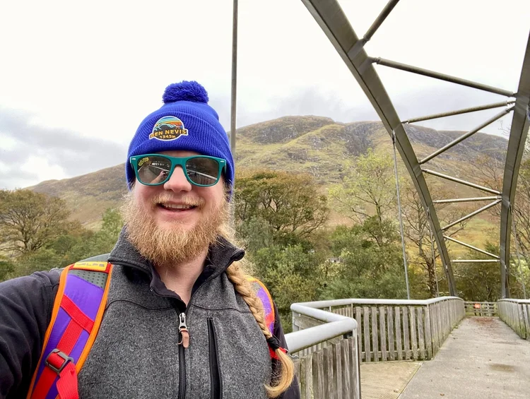 Jimmy in a blue beanie on a footbridge below the green slopes of Ben Nevis