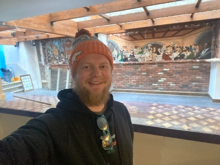 Jimmy smiling in an orange beanie inside the empty pub, a brick wall and mural behind him